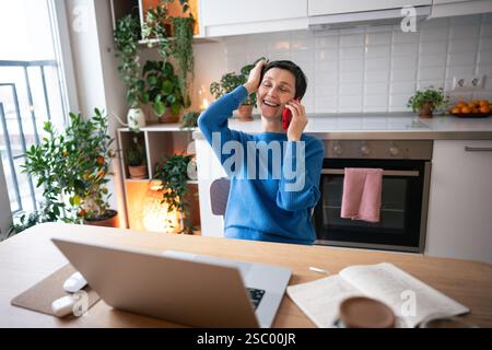 Smiling mature woman chats on phone, work on laptop and setting up online training sessions at home Stock Photo