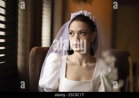 Bride looks introspective sitting by window, soft light filtering through blinds Stock Photo