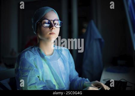 A female doctor in blue attire and glasses focused during night shift work Stock Photo