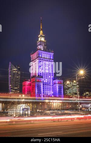 Warsaw Palace of Culture glows in red, white, and blue against the night sky, surrounded by modern skyscrapers. Light trails from cars and trams Stock Photo