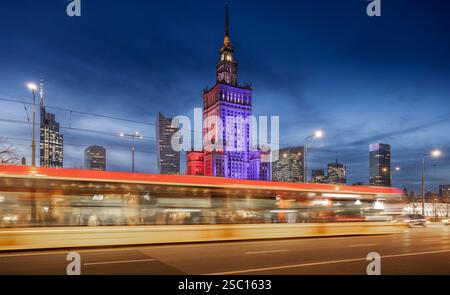 Warsaw Palace of Culture glows in red, white, and blue against the night sky, surrounded by modern skyscrapers. Light trails from cars and trams Stock Photo