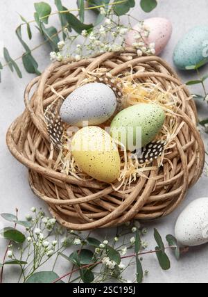 Easter eggs with pastel colors lying in wicker nest with straw and feathers, surrounded by flowers and eucalyptus leaves Stock Photo