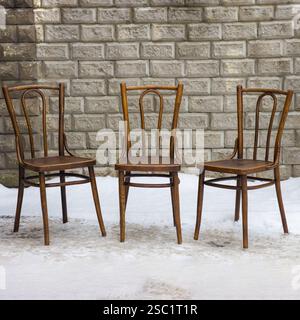 Three vintage bentwood chairs, elegantly crafted and finished in brown, stand side by side against a textured grey brick wall on a snowy surface. Stock Photo