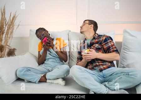 Multiethnic couple sitting on a white sofa, ignoring each other while using their smartphones, highlighting the impact of technology on modern relationships Stock Photo