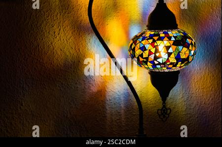 Turkish mosaic table lamp in oriental style with a colorful pattern of triangular crystals. The lamp is illuminated and casts a warm glow on the wall Stock Photo
