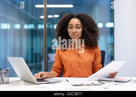 Portrait of a serious confident businesswoman doing paperwork. Financier looking at camera with documents in hands. Office worker doing paperwork inside office, using laptop. Stock Photo