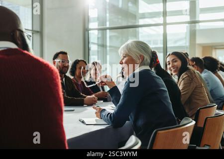 Diverse group of business professionals in a meeting with a woman speaking. Colleagues engage in a collaborative discussion in a modern conference roo Stock Photo