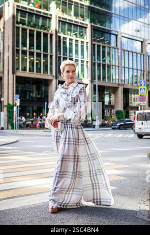 Fashionable Middle-aged woman wearing long plaid dress and stylish accessories standing confidently in urban Street near modern building, timeless fas Stock Photo