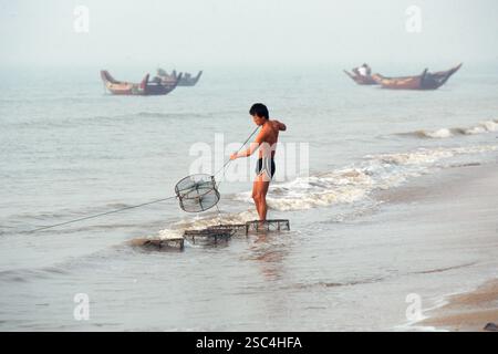 A fisherman sets a trap along a shoreline with boats visible in the background. Stock Photo