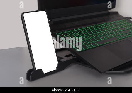 Modern gaming laptop with green backlit keyboard and a smartphone with blank screen on a holder Stock Photo