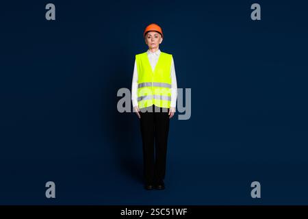 Mature businesswoman in safety gear with helmet and vest standing confidently against dark blue background. Stock Photo