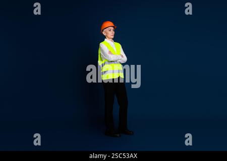 Mature female engineer standing confidently in safety gear with hardhat and reflective vest on a dark blue background Stock Photo