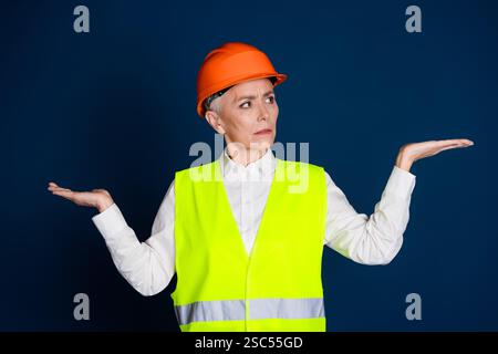 Mature female engineer in bright safety gear with thoughtful expression and arms outstretched, standing against a dark blue background. Stock Photo