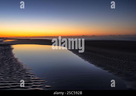 Scenic twilight over a tidal pool on a beach, reflecting golden hues of sunset against rippled sand and tranquil water, creating a serene atmosphere. Stock Photo