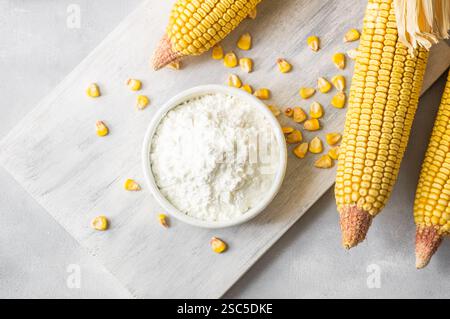 Corn starch in bowl and spoon with dried corn groats, kernels on rustic table. Maize starch with ripe cobs and kernels Stock Photo