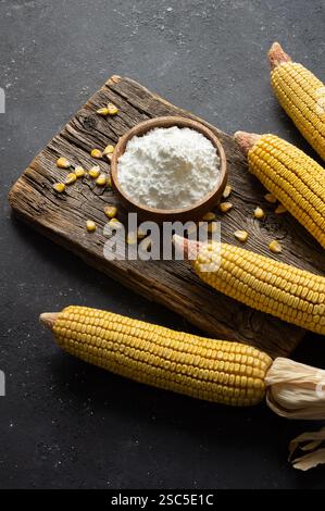 Corn starch in bowl and spoon with dried corn groats, kernels on rustic table. Maize starch with ripe cobs and kernels Stock Photo