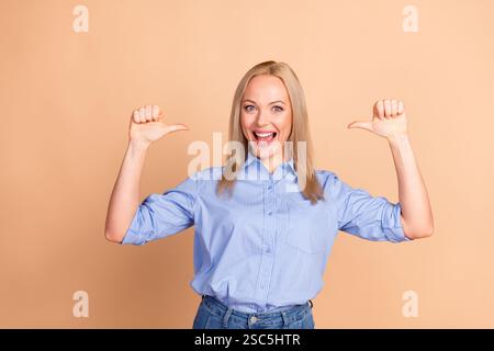 Cheerful blonde woman in a blue striped shirt posing on a beige background, joyfully pointing to herself Stock Photo