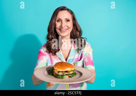 Photo portrait of pretty retired female eat hold plate burger wear trendy colorful outfit isolated on cyan color background Stock Photo