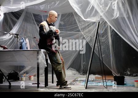 Full length shot of professional male tailor trying on custom jacket looking at reflection in mirror, while upcycling vintage clothing items into new patchwork garment in workshop Stock Photo