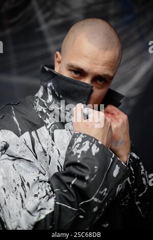Vertical portrait of male fashion designer with buzz cut confidently posing in coat customized with abstract white print, covering face with turned up collar in studio Stock Photo