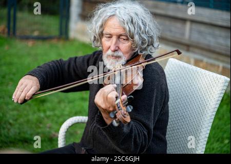A senior man with curly gray hair and a beard plays the violin while sitting on a white chair in a garden, deeply focused on his music. Stock Photo