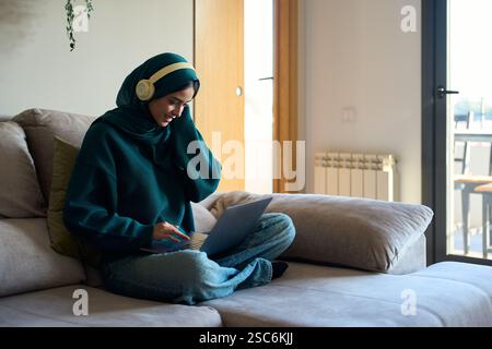 Smiling muslim woman wearing headphones using laptop on sofa at home Stock Photo
