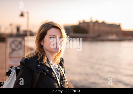 A woman stands by a waterfront at sunset, smiling with the cityscape in the background. Copenhagen, Denmark Stock Photo