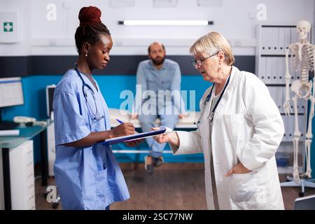 Senior doctor in lab coat discusses patient chart with black nurse, as patient sits on hospital bed in background. Elderly physician and woman in blue scrubs with clipboard standing in medical office. Stock Photo