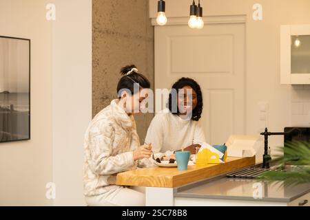 Two diverse women sharing a delightful moment, enjoying rich chocolate cake together in a bright, modern kitchen filled with warmth and laughter, cele Stock Photo