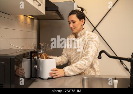 Young woman preparing toast in a modern kitchen, using a sleek white toaster to create a quick and easy breakfast or snack, enjoying a cozy morning ro Stock Photo