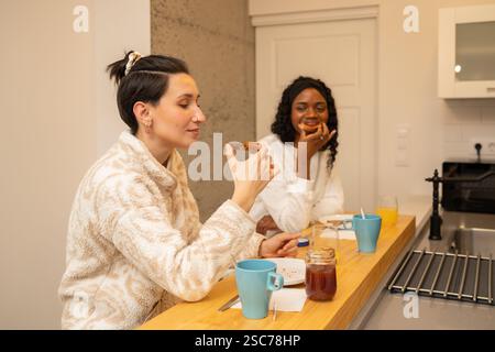 Two diverse women in bathrobes are sitting at kitchen counter, eating toast and drinking juice and coffee, having a pleasant morning conversation Stock Photo