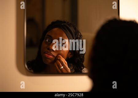 Young woman with curly hair, wearing headband, touching face while examining skin closely in bathroom mirror during nighttime skincare routine, checki Stock Photo