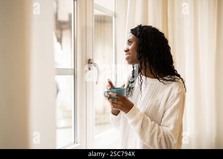 Young woman with long, curly hair holds a blue mug and gazes thoughtfully out a bright window, enjoying a quiet moment of reflection with her morning Stock Photo