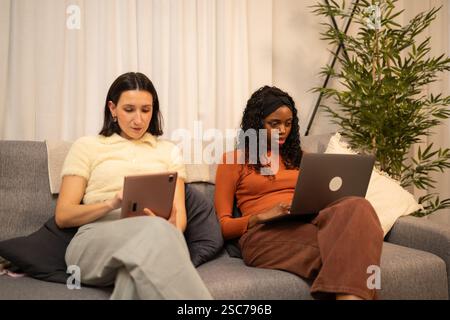 Two young women are sitting on a couch, working remotely, one using a laptop and the other using a tablet, focused on their tasks in a comfortable hom Stock Photo