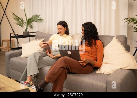 Two young women, possibly roommates or friends, are relaxing on a comfortable sofa in their modern living room, using a laptop and tablet, enjoying th Stock Photo