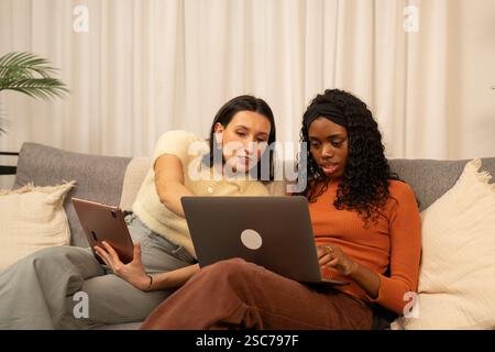 Two young women entrepreneurs are sitting on a comfortable sofa, collaborating on a project using a laptop and a tablet, discussing ideas and strategi Stock Photo