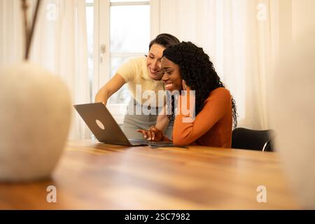 Two smiling female colleagues are collaborating on a project, using a laptop at a wooden table in a brightly lit home office, enjoying their productiv Stock Photo