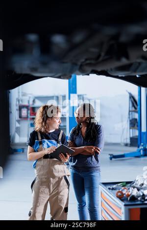 Serviceman in garage using tablet to show BIPOC client procedures checklist while doing maintenance on automobile. Car service worker does inspection on vehicle next to customer Stock Photo