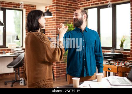 Businessman and woman in startup company high five near desk, displaying successful collaboration. Young female and male employees celebrating good teamwork and project presentation in office. Stock Photo