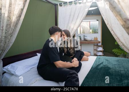 Young couple holding hands and enjoying an intimate moment together while sitting on bed inside their glamping tent, showcasing romance and unique tra Stock Photo
