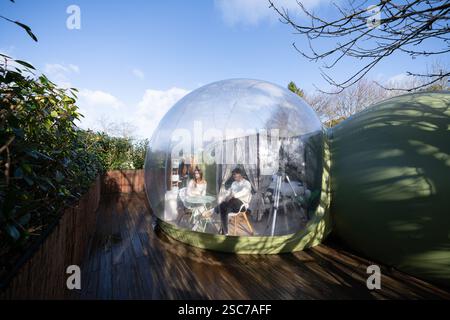 Young couple enjoying their stay in a unique, transparent bubble hotel room, providing a panoramic view of the surrounding nature, sitting at a small Stock Photo