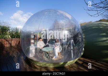 Couple enjoying a glamping experience, relaxing inside a transparent inflatable bubble tent on a wooden terrace, offering panoramic views of the sky Stock Photo