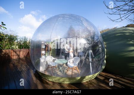 Romantic couple embracing inside a transparent bubble hotel room, featuring furniture and curtains on a wooden deck beneath a clear blue sky, creating Stock Photo