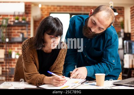 Female employee holding clipboard and planning business project presentation with assistance from manager. Caucasian coworkers leaning on desk and collaborating on strategy ideas for startup company Stock Photo