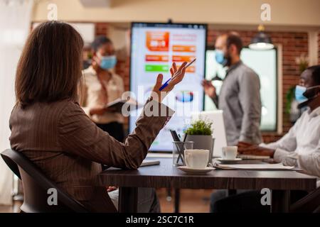 Multiethnic team wearing face masks for safety in office and discussing strategy ideas with information seen on monitor. Closeup on woman with raised hand, holding a pen and speaking to her coworkers. Stock Photo