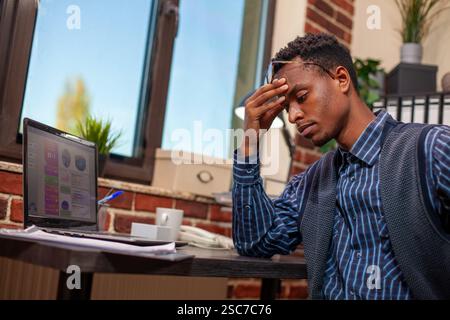 African American entrepreneur sits tiredly at desk, holding his head as he analyzes business project plans. Black male manager looking drained after demanding day at work in startup office. Stock Photo