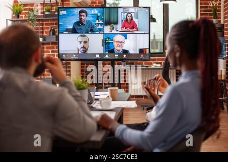 Workers in office discussing strategy ideas with colleagues through a virtual meeting platform on screen. Entrepreneurs participate in online call, collaborating on marketing plan in video webinar. Stock Photo