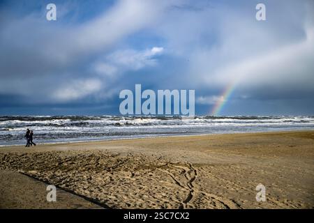 A couple walks along a sandy beach with a rainbow in the sky and waves crashing on the shore. Stock Photo