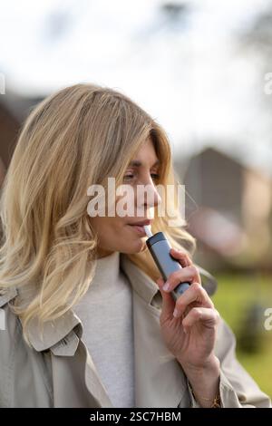 A woman with long blonde hair takes a slow puff from a vape device while standing outdoors. The background features a tranquil neighborhood environmen Stock Photo