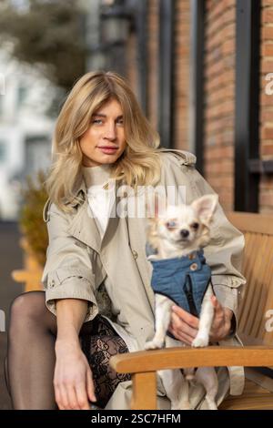 A woman with long blonde hair sits on a wooden bench holding her small dog dressed in a blue outfit. They are in an urban area with brick buildings in Stock Photo